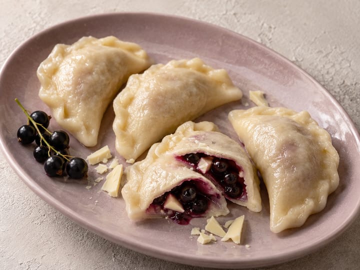 Pierogi fantazyjne na liliowym półmisku; w rozciętym pierogu czarna porzeczka i biała czekolada, obok gałązka porzeczek