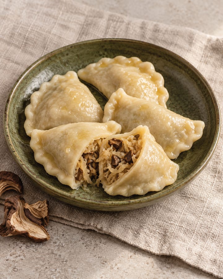 Pierogi z kapustą kiszoną i grzybami na zielonym talerzu; rozerwany pieróg pokazuje farsz z kiszonej kapusty i grzybów, obok suszone grzyby